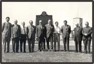 Photo en noir et blanc de 10 hommes en habits posant devant une plaque