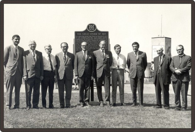 Black and white photo of 10 men in suits in front of a plaque.