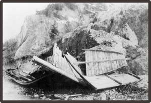Photo en noir et blanc d’un bateau échoué sur une plage s’étant brisé contre des rochers.