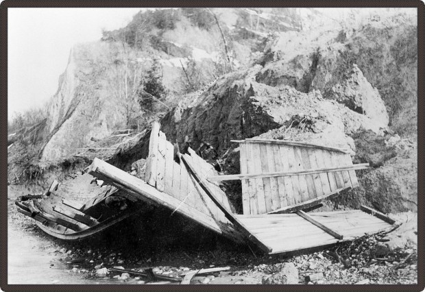 Black and white photo of a boat washed up on a beach, shattered by the rocks.