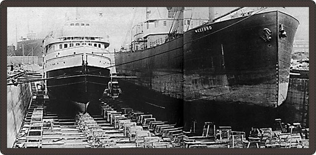 Black and white photo of two boats on dry land. "Wexford" is written on the side of the bigger boat.