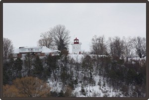 Photo of a winter landscape with a lighthouse in the distance, on top of a hill next to a big house.