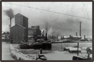 Black and white photo of a snow-covered port and factory. Three boats are docked at harbour with pieces of ice floating around them.