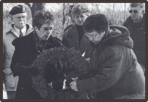 Photo en noir et blanc de deux femmes déposant une couronne de fleur dans un cimetière. Derrière eux se trouvent quatre personnes.