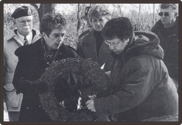 Black and white photo of two women putting down a wreath in a cemetery with four people behind them.