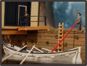 Wooden model of a small four-paddle rowboat with small wooden models of people for scale.