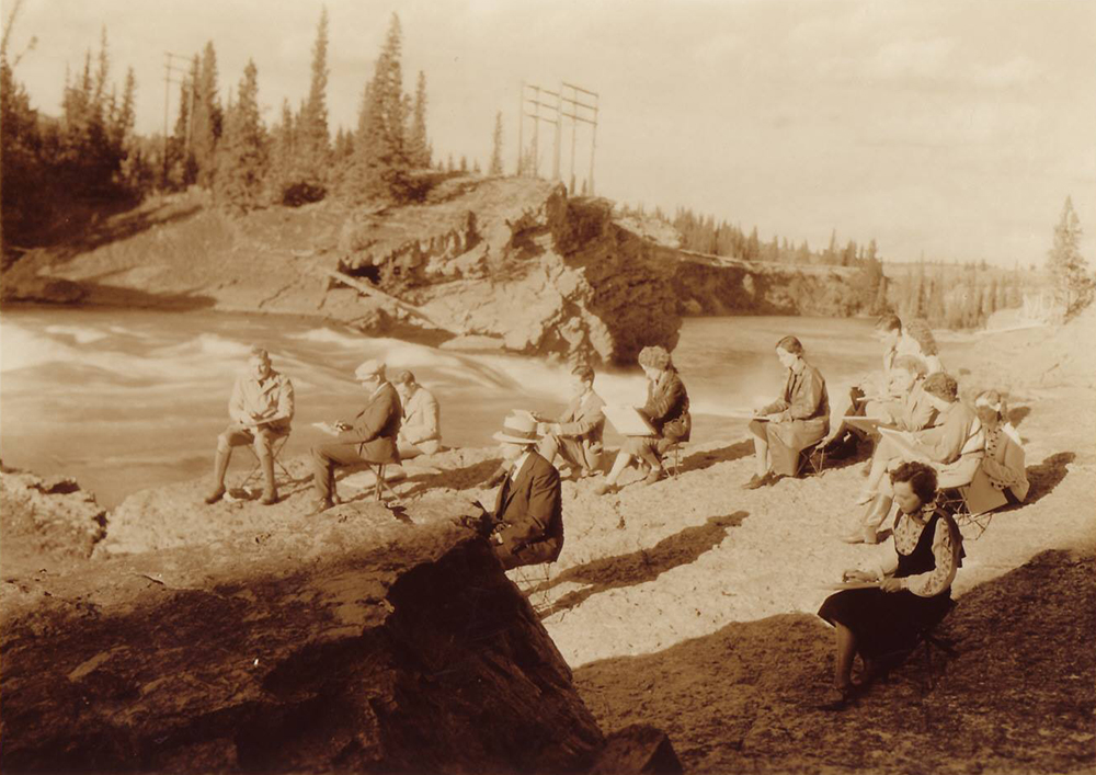 Sepia photo of group of artists sketching on bank of river in treed and rocky landscape.