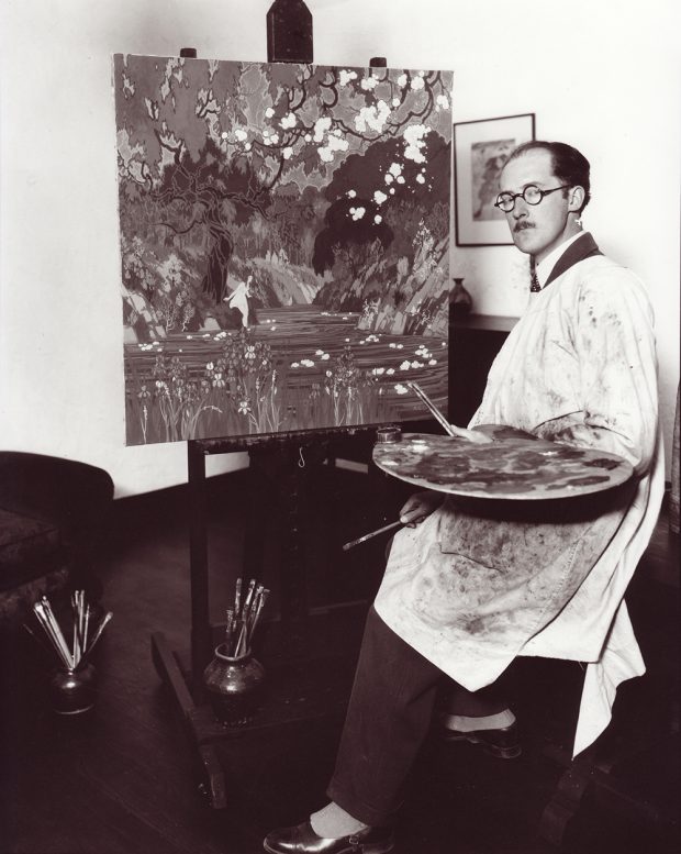 Black and white photo of man in white smock, holding paintbrushes and palette, sitting next to easel with painting of girl next to river.