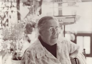 Black and white photo of woman with grey hair and glasses seated in bright room with blurry background.