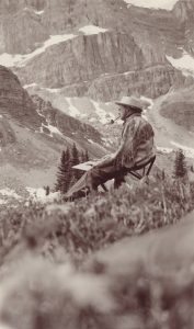Black and white photo of man wearing fringed buckskin jacket, hat and smoking pipe, painting in a mountain landscape.