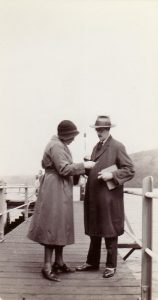 Black and white photo of sharply dressed couple standing on dock, consulting something in woman's hand.