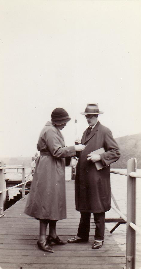 Black and white photo of sharply dressed couple standing on dock, consulting something in woman's hand.