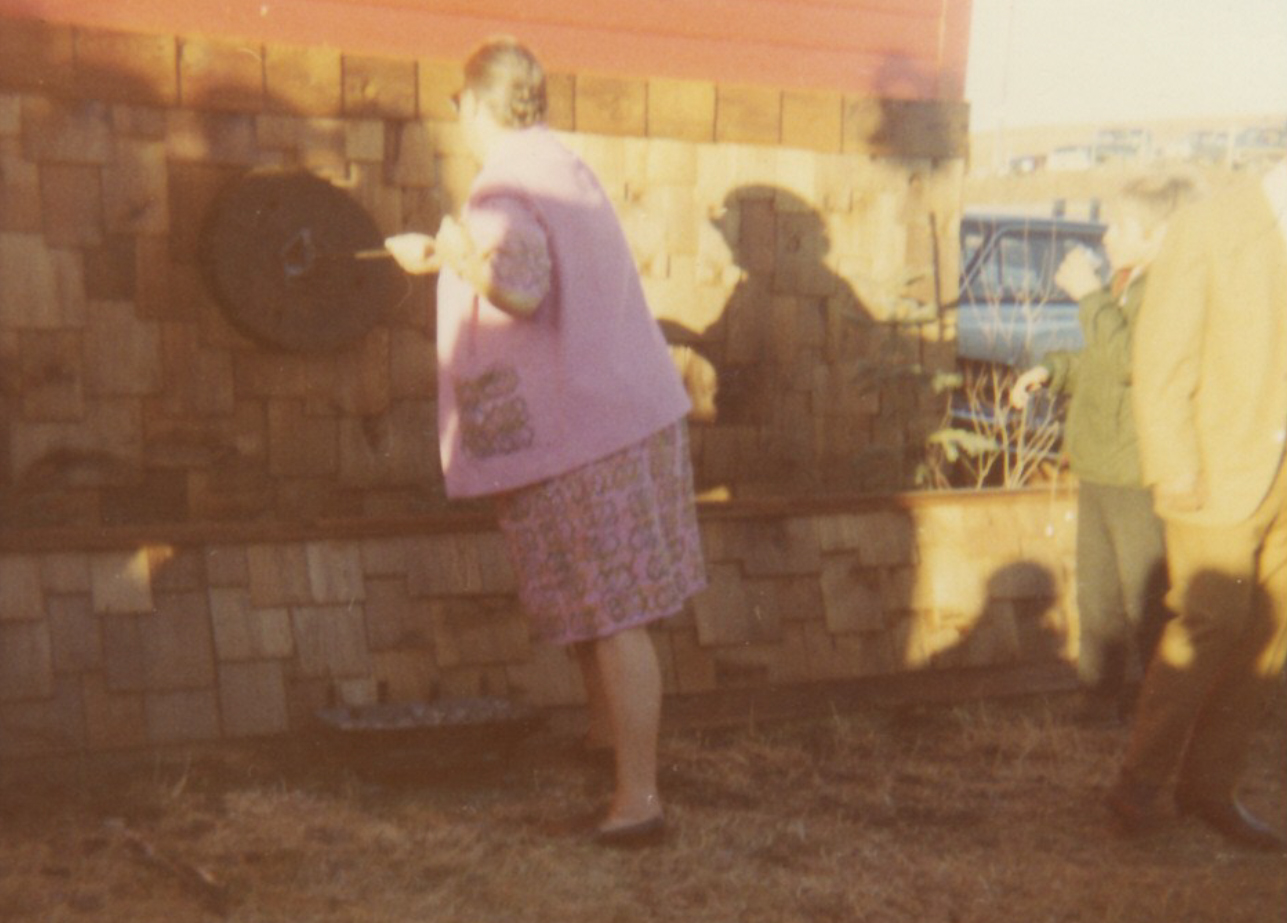 Colour photo of woman facing away, with branding iron in hand, branding circular object on outdoor wall.