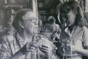 Black and white photo of woman weaving on cardboard loom with young girl.