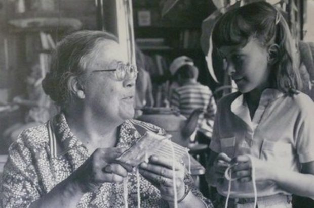 Black and white photo of woman weaving on cardboard loom with young girl.