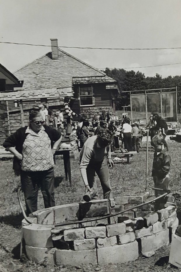 Black and white photo of two adults and one child around fire pit with old fashioned schoolhouse behind.