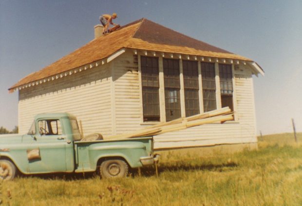 Colour photo of man roofing an old-fashioned white schoolhouse with green pickup-truck in front.