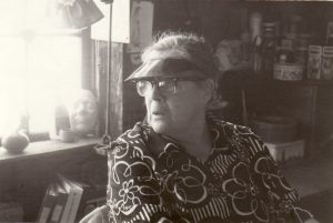 Black and white photo of woman wearing visor and glasses with tins, binders and knick knacks in background.