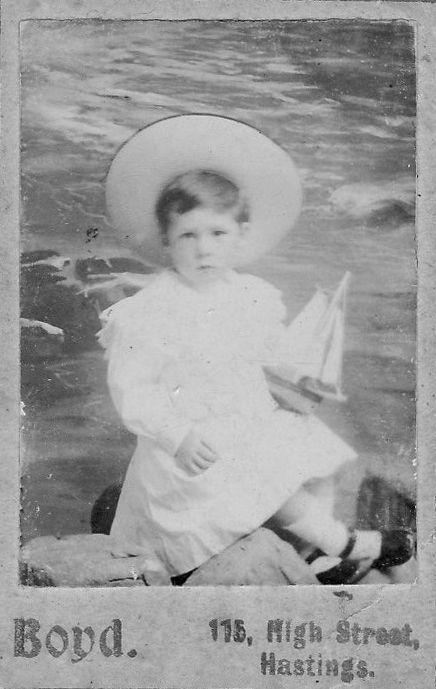 Black and white studio portrait of young boy holding toy boat and wearing large hat and white dress.