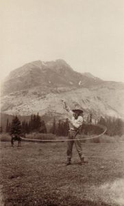 Sepia photo of man in hat with lasso standing in front of mountain and horse in distance.