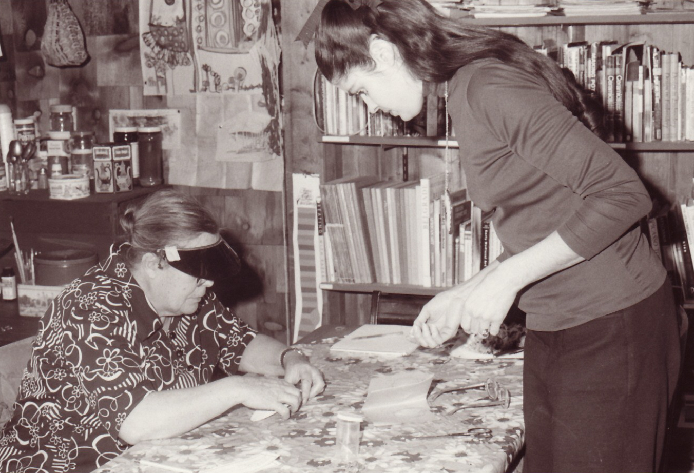 Black and white photo of woman working at a table with younger woman student standing opposite; bookshelf and art supplies behind.