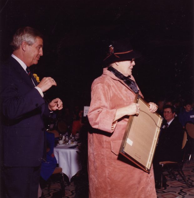 Colour photograph with a woman wearing a hat and suede jacket, holding a frame; man in a suit stands behind her and a crowd of people seated at tables look on.