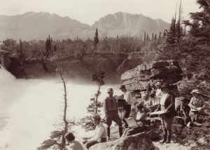 Sepia photo of group of artists sketching on bank overlooking a waterfall in treed and mountain landscape.