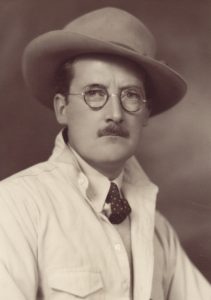 Sepia studio portrait of man with moustache wearing glasses, felt hat, tie and light coloured jacket.