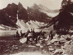 Sepia photo of a group of people riding horses next to a lake in a mountain landscape.