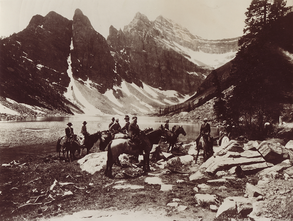 Sepia photo of a group of people riding horses next to a lake in a mountain landscape.