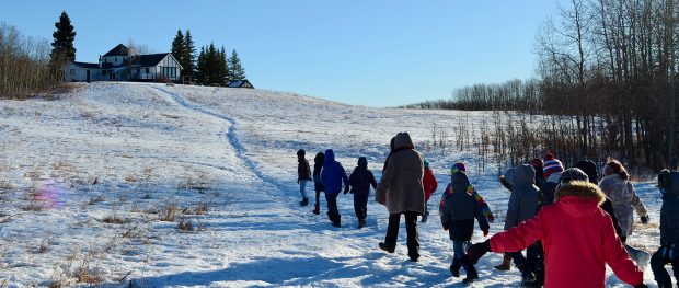 Colour photo of a line of children walking single file up a hill in a winter landscape towards a half timbered house.