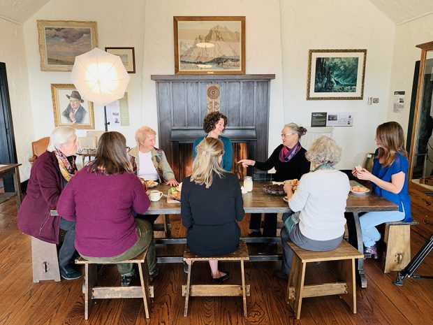 Colour photograph of group of women eating around table; artwork on walls in background.