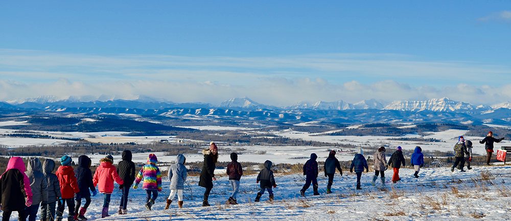 Colour photo of a line of children walking single file in front of a winter panorama of mountains in the distance.