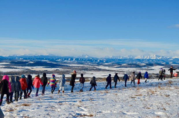 Colour photo of a line of children walking single file in front of a winter panorama of mountains in the distance.