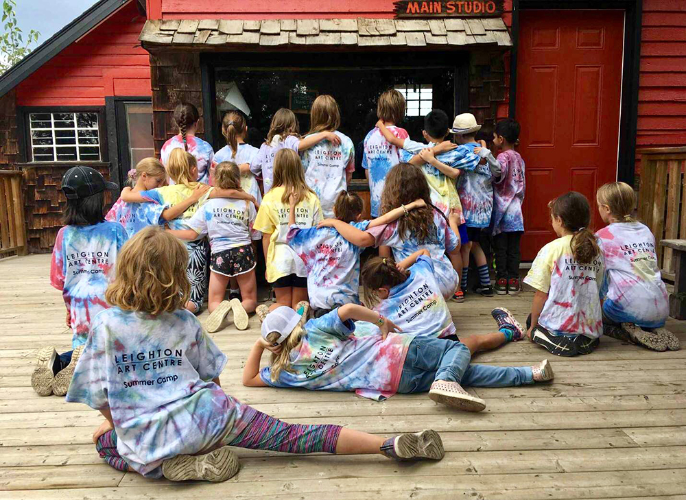 Colour photograph of group of children on a deck, wearing tie-dyed t-shirts and facing a red building.