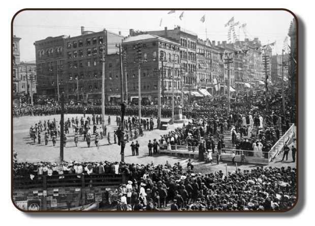 A black and white image of what a city-wide celebration in downtown Saint John, New Brunswick would have looked like in 1867, upon the arrival of The Paris Crew when they returned from Paris. The intersection of King Street and Water Street are lined with thousands of people. In the opened central square area, celebration activities are being performed for the spectators. A make-shift, multi-level stadium seating area was also erected directly in front of the square. Decorations adorn the store fronts and flags line the telephone poles and lamp posts.
