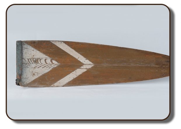 An image of a wooden oar that was reported used by the members of The Paris Crew. At the tip of the paddle end of the oar is a decorative triangle shaped paint detail in white. There is also a leather covering over the blade, which is damaged and pealing away on one end either by repetitive use or age.
