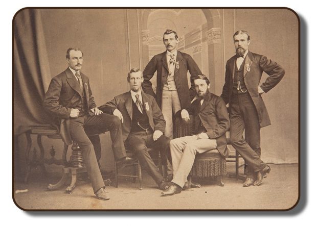 A sepia toned studio photograph of The Paris Crew. All four members and their manager Sherriff J. A. Harding are present in the photograph. The men are all positioned together sitting upon different types of antique furniture. All are wearing formal suit and tie attire, as this photograph was taken as part of the celebrations that took place upon their return from Paris in 1867.