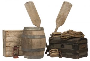 Decorative image of a collage of materials that would have been seen at a dock or pier at the time of The Paris Crew in 1967. The collage includes a wooden barrel with four metal support rings, a wooden trunk, a red kerosene lantern, a bundle of fisherman's rope, two pulleys that would have been used to crank either the sails or mast on a ship, a light birch wood create and two wooden oars.