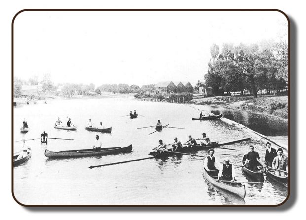 Image of a riverbank in the Renforth area of Rothesay, New Brunswick prior to the Paris Crew's participation in the International Rowing Regatta in July 1867. This photograph is in black and white, and quite faded due to its age and personal photography equipment available at that time. There are a number of different boats on the water including the Paris Crew in their four-person rowing scull, single rowers, and others in one-person and two-person canoes. Along the banks of the river is a wooded area and what appear to be three barns or boat houses.