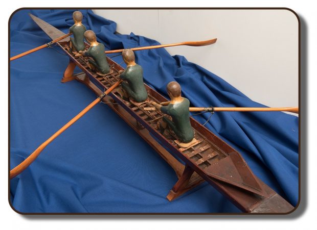 Image of a wooden replica of the famed J. A. Harding rowing scull that the Paris Crew would have competed with at the International Rowing Regatta in Paris, France. This model is approximately three feet in length, with four wooden figures representing the four team members.