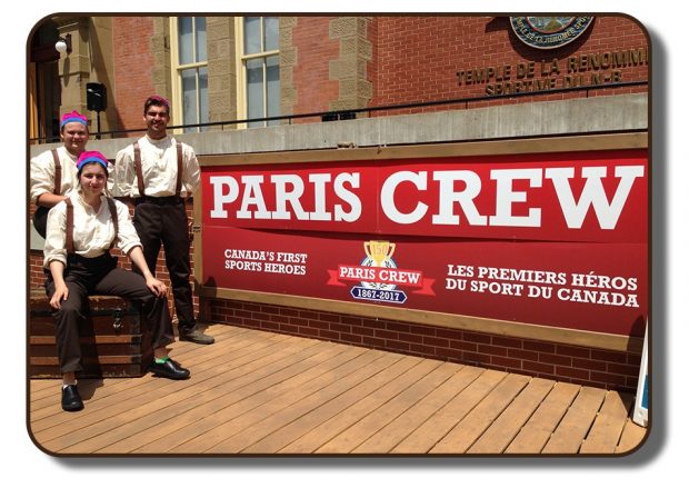 Image of an outdoor stage area in front of the New Brunswick Sports Hall of Fame. The stage is meant to resemble a wooden dock from that time period. Three performers are dressed in period clothing including brown pants, white linen shirts, brown leather suspenders and wearing the Paris Crew signature rowing caps. The backdrop of the stage area reads The Paris Crew: Canada's First Sports Heroes with a one hundred and fiftieth anniversary crest.