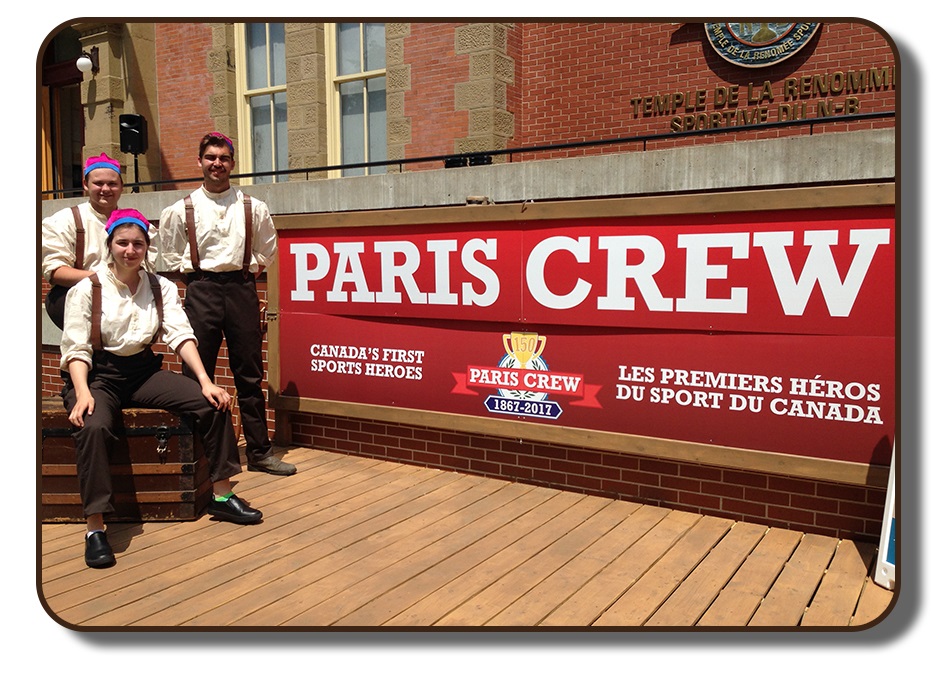 Image of an outdoor stage area in front of the New Brunswick Sports Hall of Fame. The stage is meant to resemble a wooden dock from that time period. Three performers are dressed in period clothing including brown pants, white linen shirts, brown leather suspenders and wearing the Paris Crew signature rowing caps. The backdrop of the stage area reads 