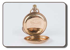 Image of a gold-coloured pocket watch with front and back hinged covers opened up to show the engaged markings on the back. The engraving shows that this particular pocket watch was presented to Samuel Hutton, member of the Paris Crew for rowing in 1867. The other hinged cover was used to protect the face of the watch, which is not visible from the angle shown.