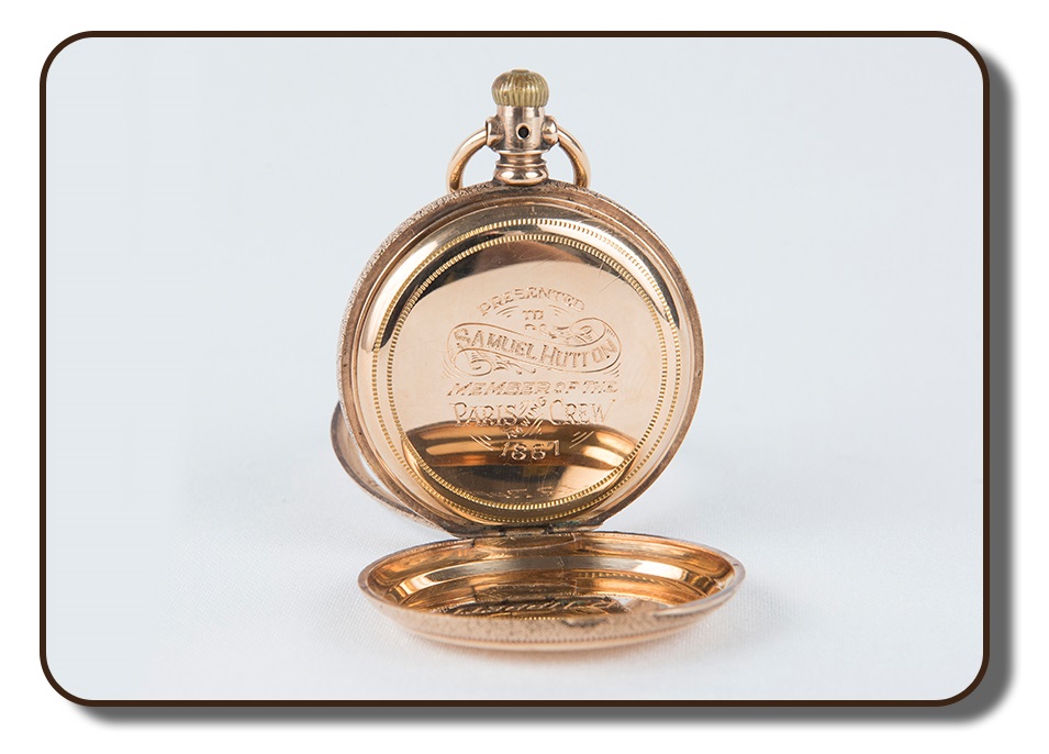 Image of a gold-coloured pocket watch with front and back hinged covers opened up to show the engaged markings on the back. The engraving shows that this particular pocket watch was presented to Samuel Hutton, member of the Paris Crew for rowing in 1867. The other hinged cover was used to protect the face of the watch, which is not visible from the angle shown.