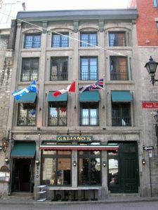 Colour photograph showing the façade of a four-storey building, with green shutters adorning some windows. The flags of Quebec, Canada and the United Kingdom, hung from the second floor, flutter in the breeze.