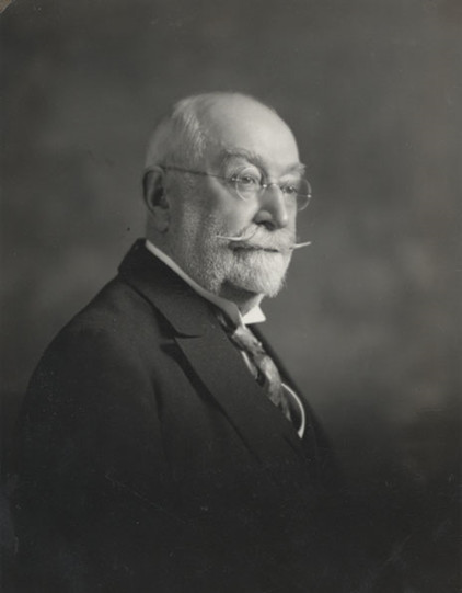 Black & white photograph of an older man with a short white beard and a long upturned moustache. He wears round eyeglasses, a white shirt with a tie, and a black jacket.