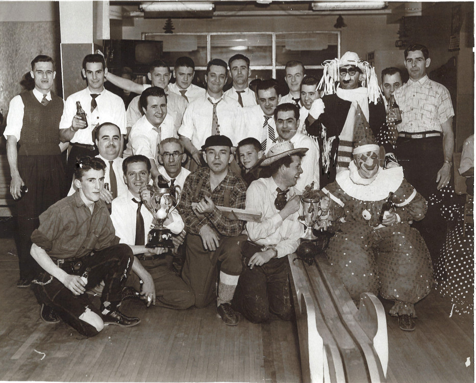 Black & white photograph of a group of men, some in costume, next to a bowling alley.