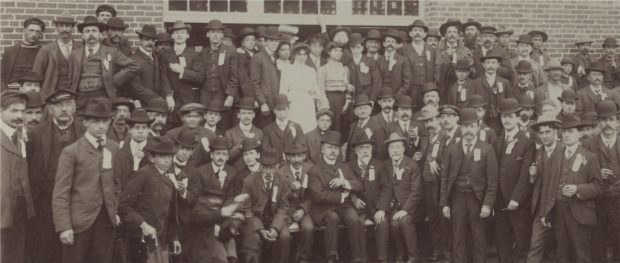 Sepia-tone photograph showing men and some women outside, in front of a paper mill.