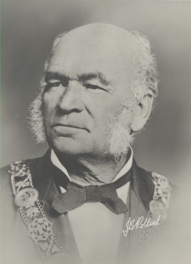 Portrait in black & white of an elderly man with white sideburns. He wears a white shirt with a black bow tie, and a dark jacket with ornamentation (a chain of office).